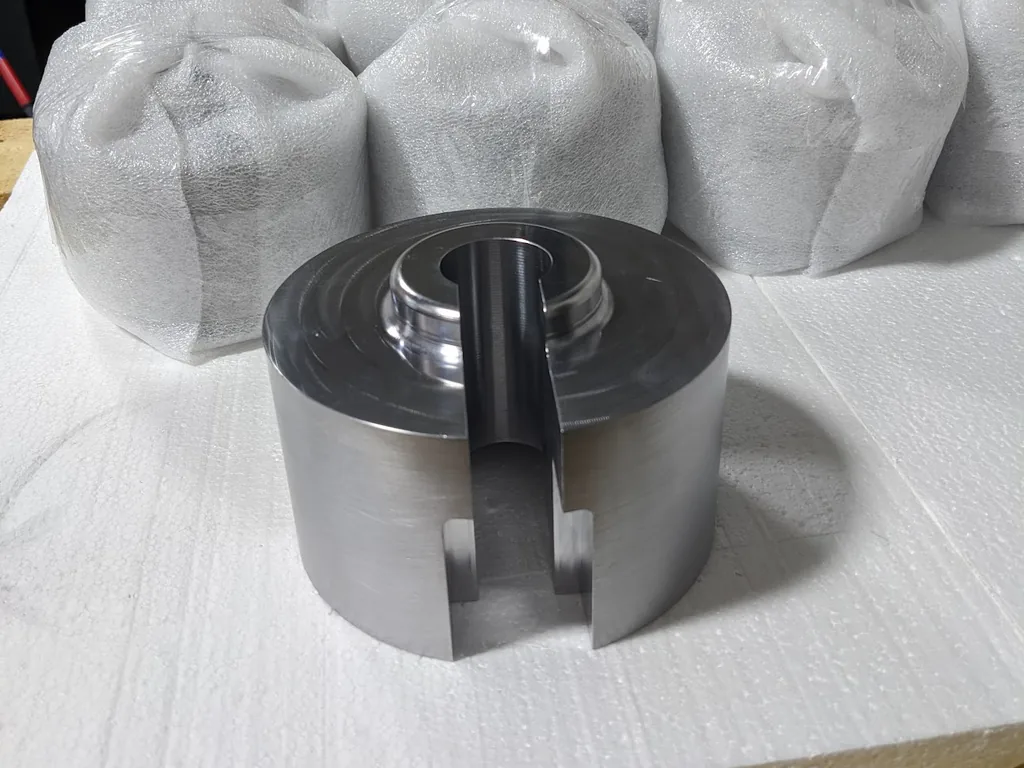 Large turned metal fixture with a center slot on white foam, with bubble-wrapped parts behind it ready to ship